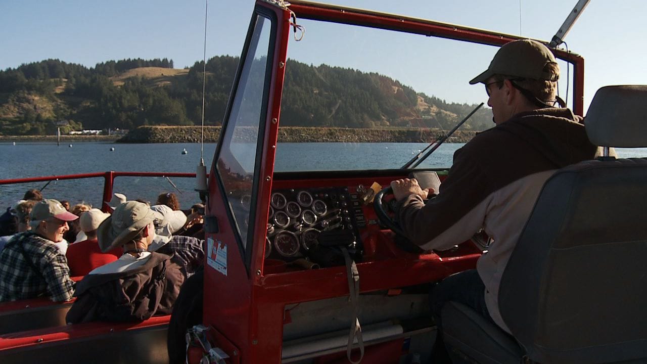 A person drives a red tour boat with passengers seated, against a scenic backdrop of a lake and forested hills under a clear blue sky.