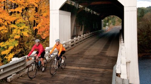 Cyclists pedal through covered bridge and past fall foliage.