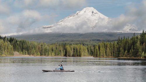 A kayaker paddles on a glassy lake with Mt. Hood's peak in the background.