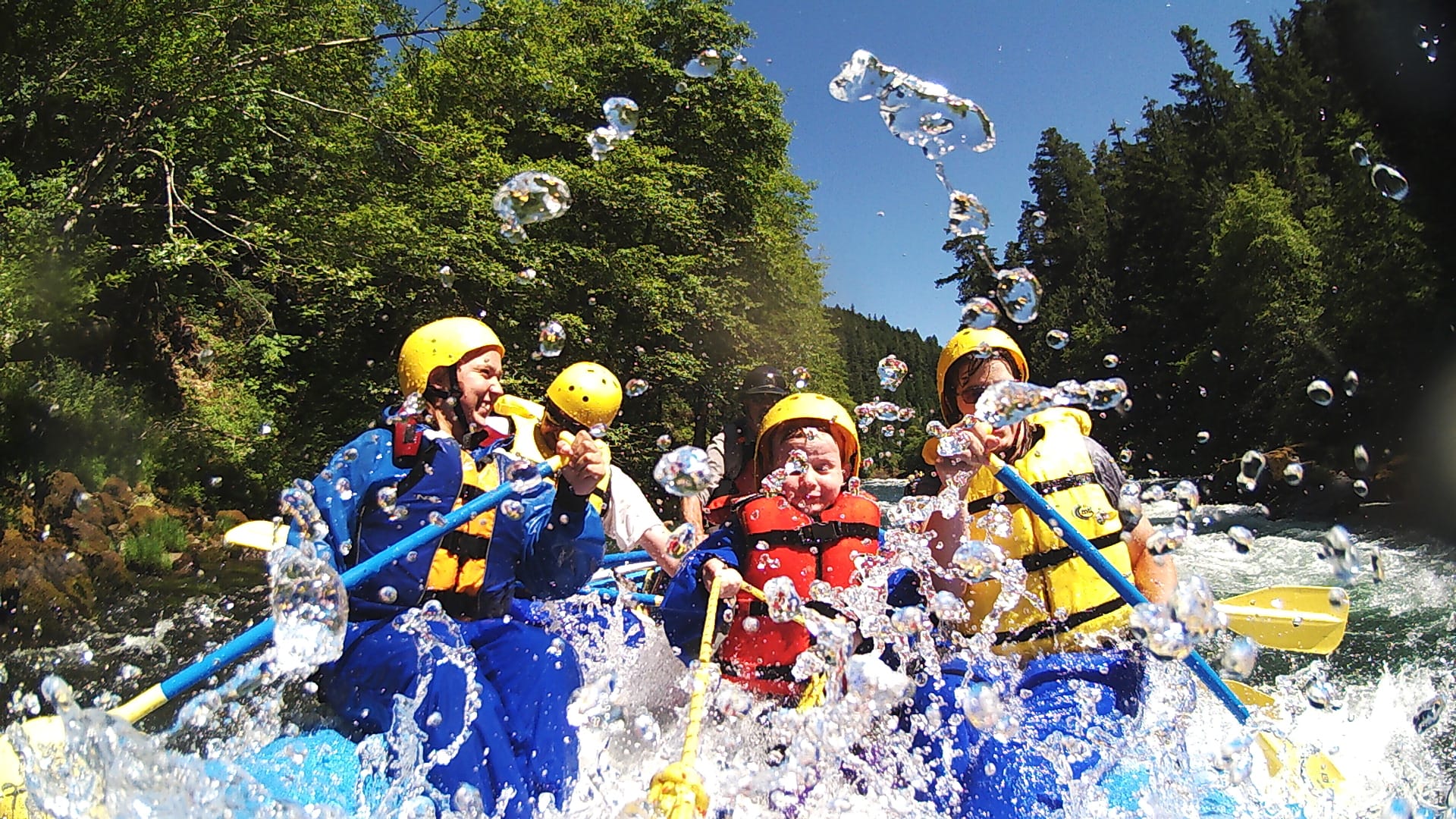 People in colorful life jackets and helmets paddle a raft, navigating through splashing rapids surrounded by lush green trees and clear blue skies.