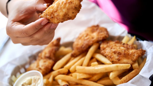 Hand holding fried chicken tender above a basket of french fries and more chicken tenders, with a cup of dipping sauce on the side, against a pink background.