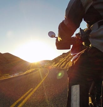 A motorcyclist rides on a curved road at sunrise, surrounded by hills. The scene captures the bright sun and clear sky, illuminating the path ahead.