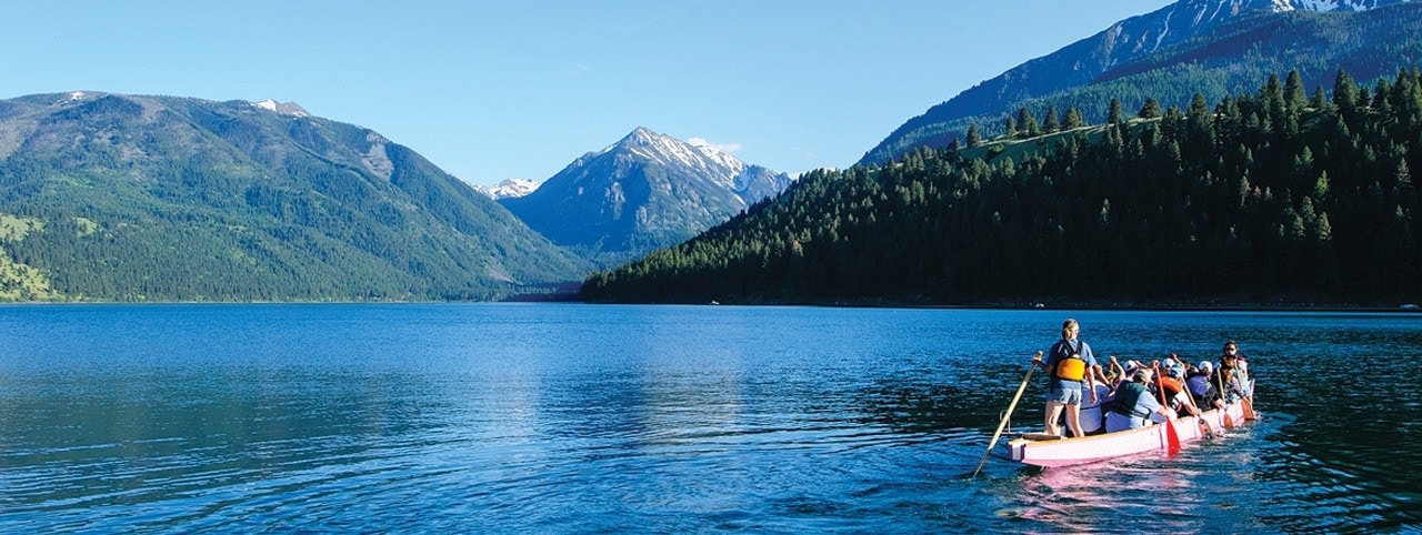 A group of people paddle a long canoe across a tranquil, blue mountain lake, surrounded by forested hills and distant snow-capped peaks under a clear sky.