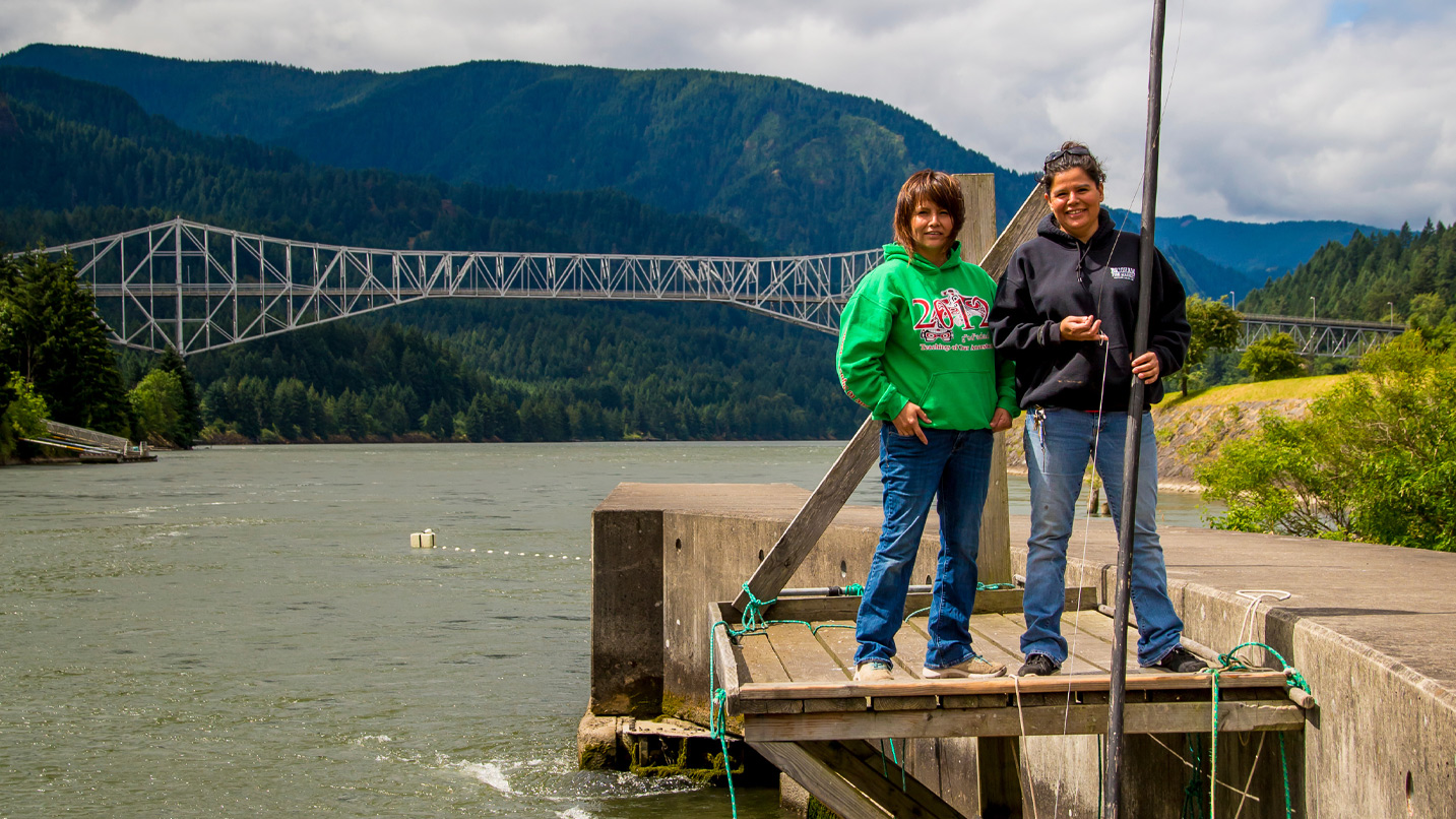 Two Native American women stand on a wooden platform over a river with a bridge in the background