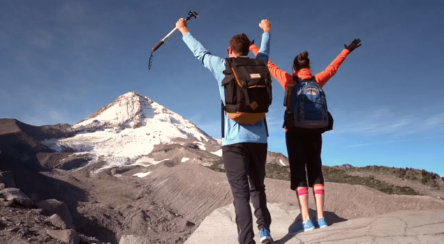 Two hikers, with arms raised triumphantly, stand on a rocky ledge, holding an ice axe. Snow-covered mountain peak dominates the background under a clear blue sky.