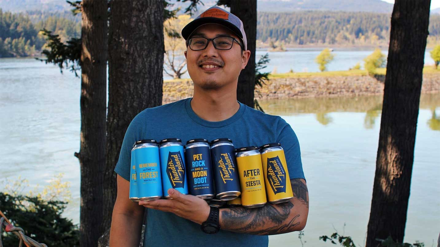 A brewer holds cans of Thunder Island Brewing in front of the Columbia River.