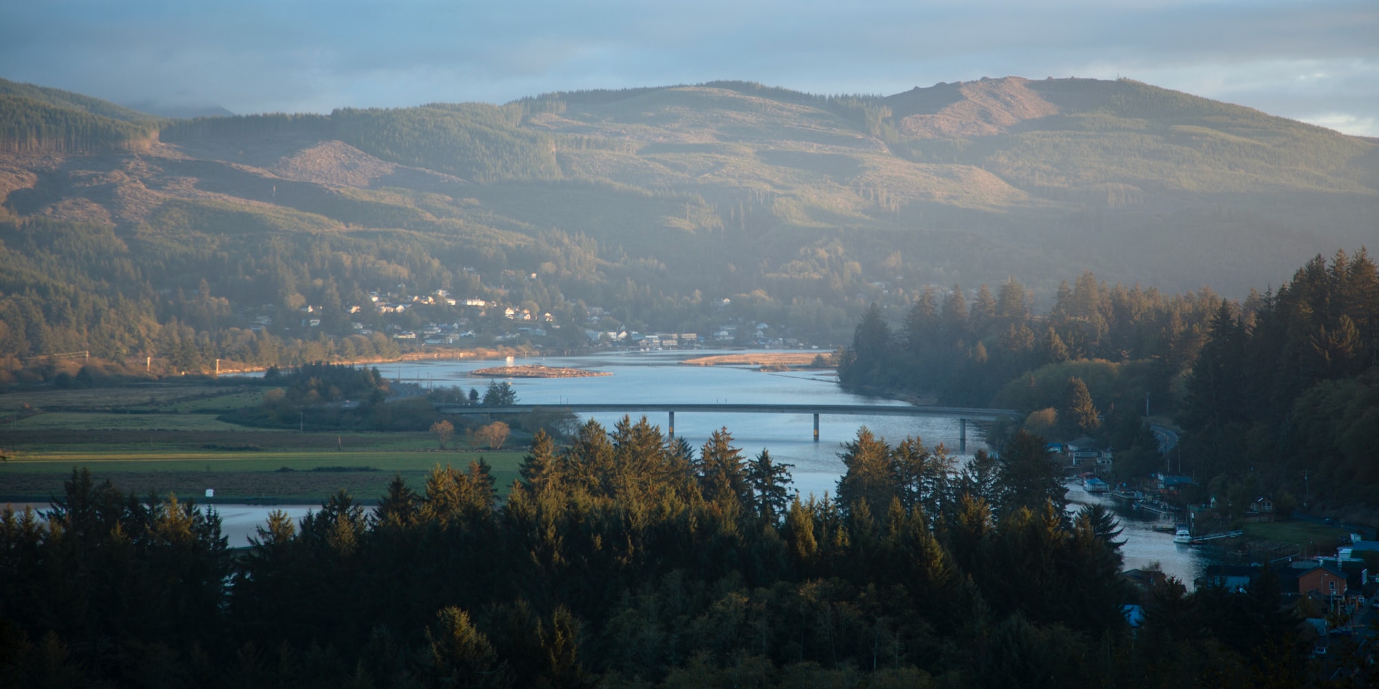 River winds through lush forest and fields, bordered by distant hills and a small town under soft light, with a bridge crossing over in the midground.