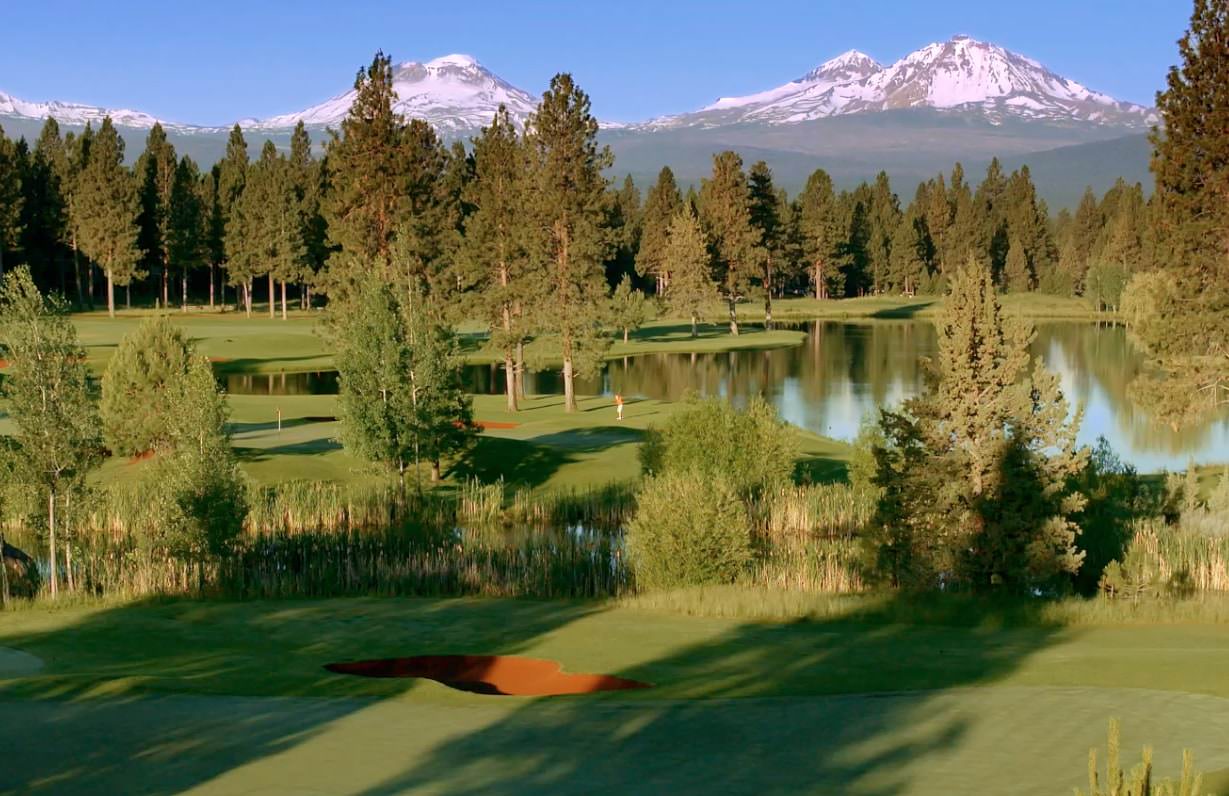 A lush golf course features a sand trap and flagpole. Trees surround a serene pond, with snow-capped mountains rising prominently in the clear, blue sky background.