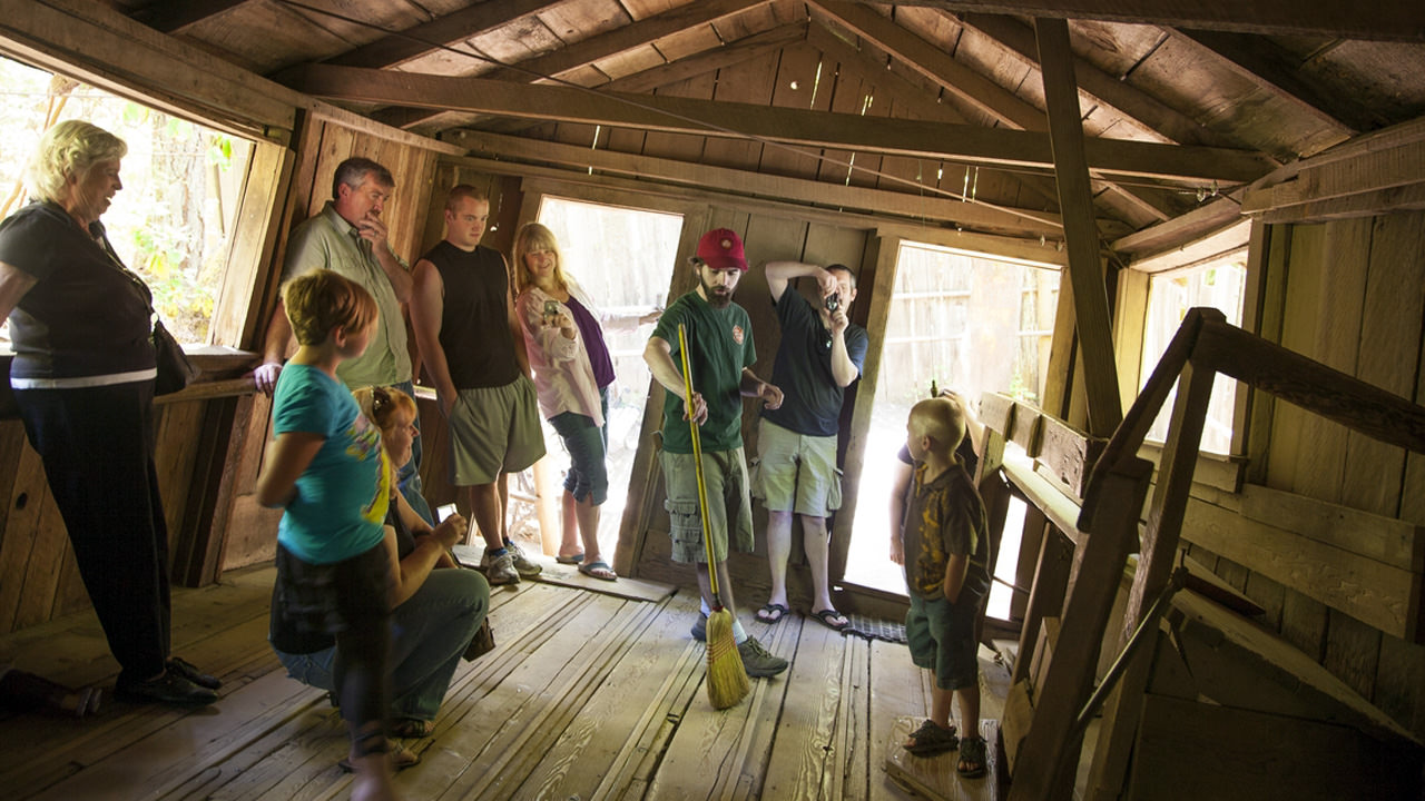 People stand in a tilted house while a volunteer demonstrates a broom standing upright