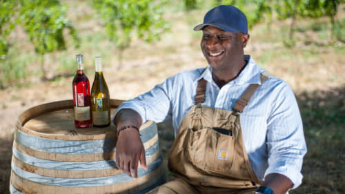 Bertony Faustin wears overalls and a collared shirt as he smiles next to two bottles of wine he made.