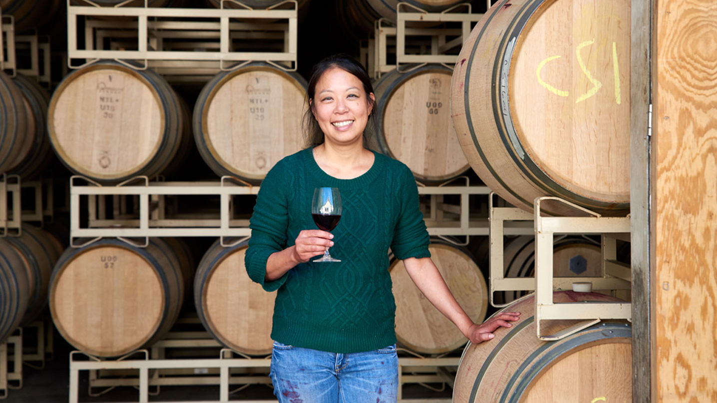 A woman holds a glass of wine in front of wine barrels
