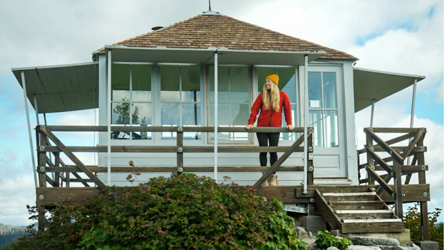 A woman in a red outdoor jacket standing on the porch of a rustic fire lookout.