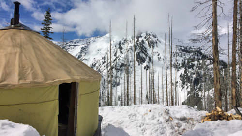 A yurt sits in the sunshine surrounded by snow.