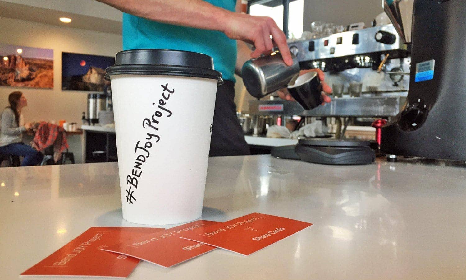 Coffee cup labeled #BendJOYProject near branded orange cards and barista