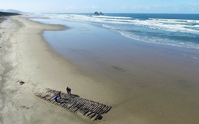 Two people stand on a wide beach near partially buried wooden beams. Waves crash in the distance, and rock formations are visible offshore under a clear blue sky.