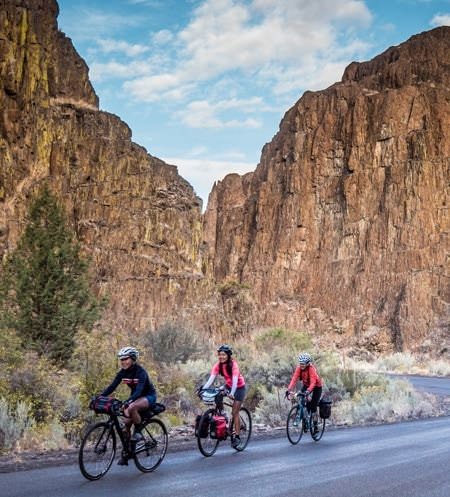Three women cyclists with gear strapped to the bikes ride through deep canyon walls