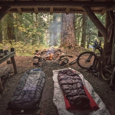 Two sleeping bags lay in an open shelter with a campfire out front