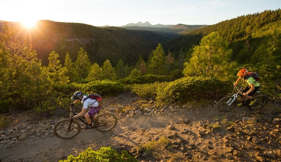 Two cyclists on a dirt trail with the sun rising above mountains