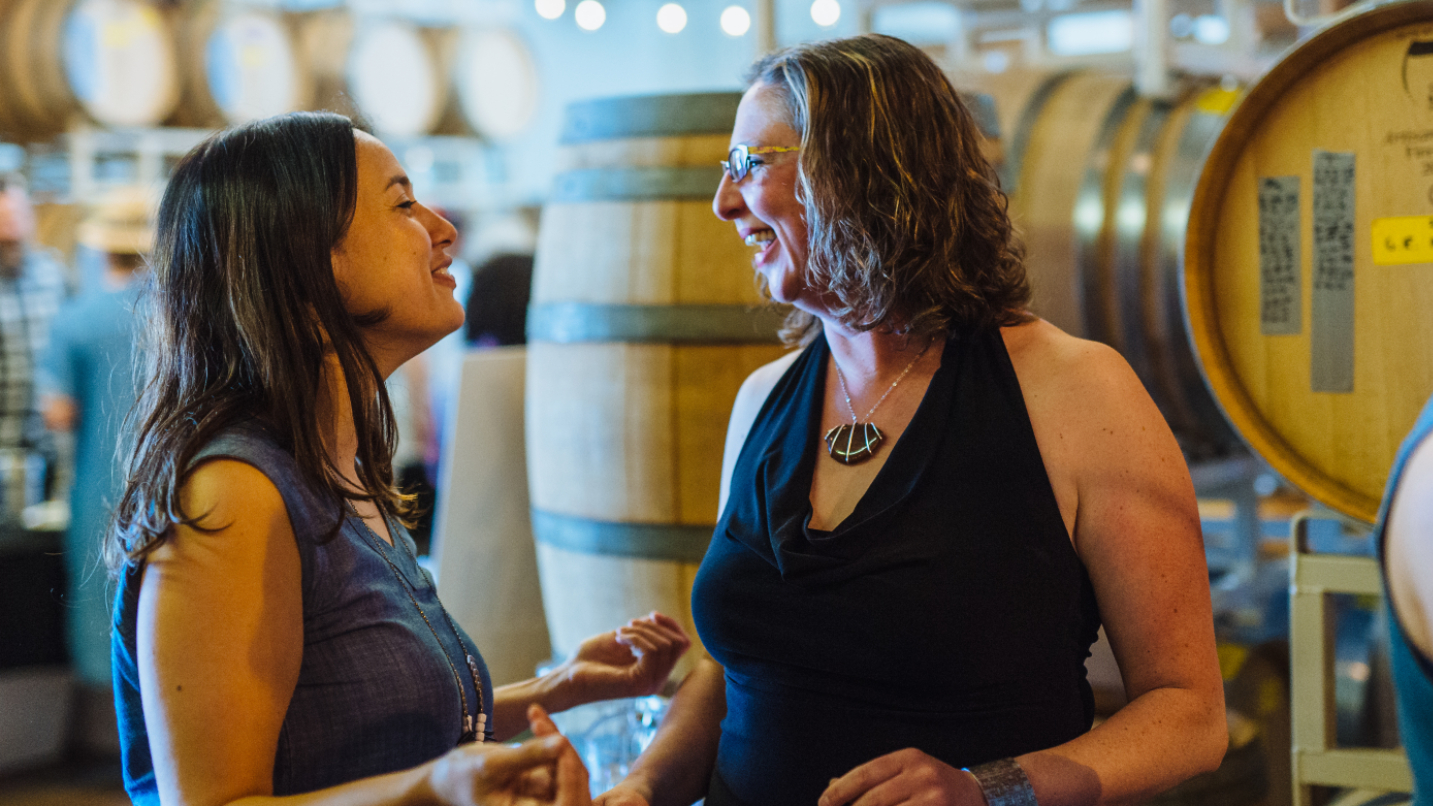 two women talk to each other with wine barrels in the background