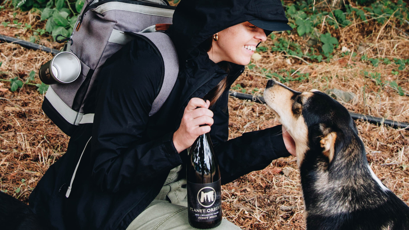 woman kneels in the woods while holds wine bottle and pets dog