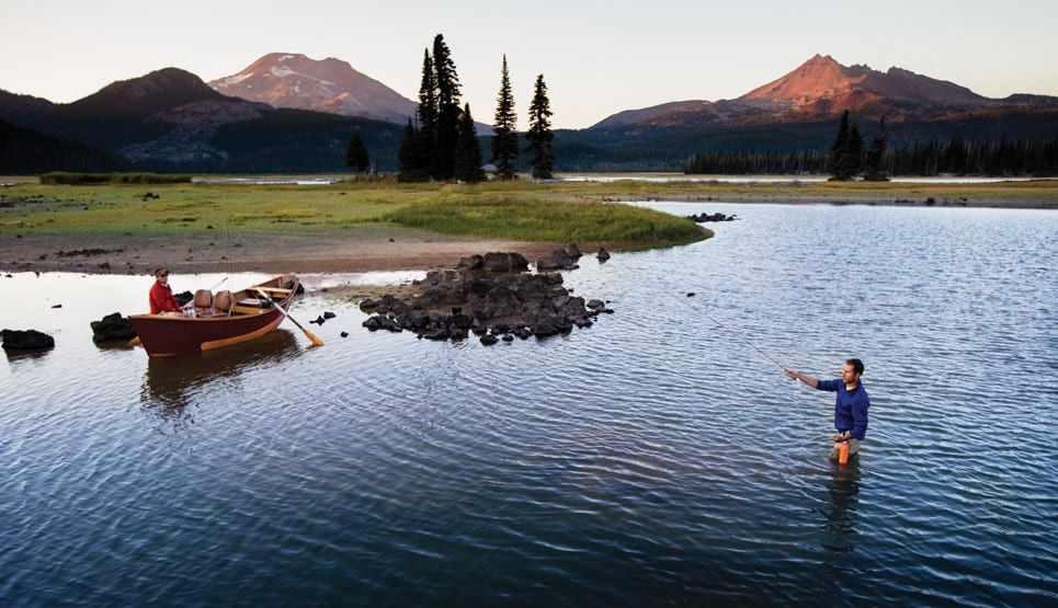 A person standing in a lake fly-fishing at sunset with mountains in the background
