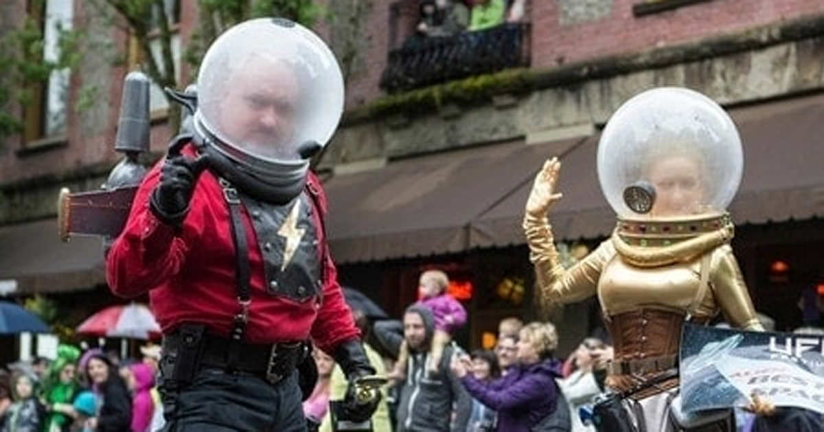 Two people dressed as retro-style astronauts with glass helmets walk in a parade. The man wears red with a lightning bolt. The woman wears gold. Onlookers and buildings fill the background.