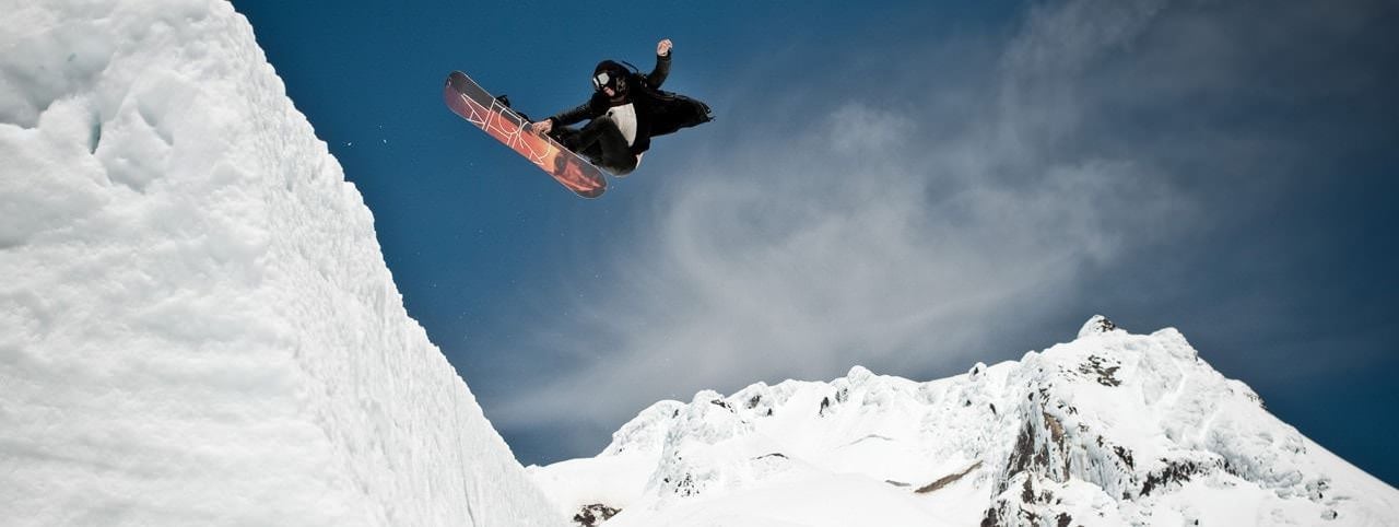 A snowboarder leaps in mid-air off a snowy ramp, against a backdrop of snow-covered mountains and a clear blue sky.