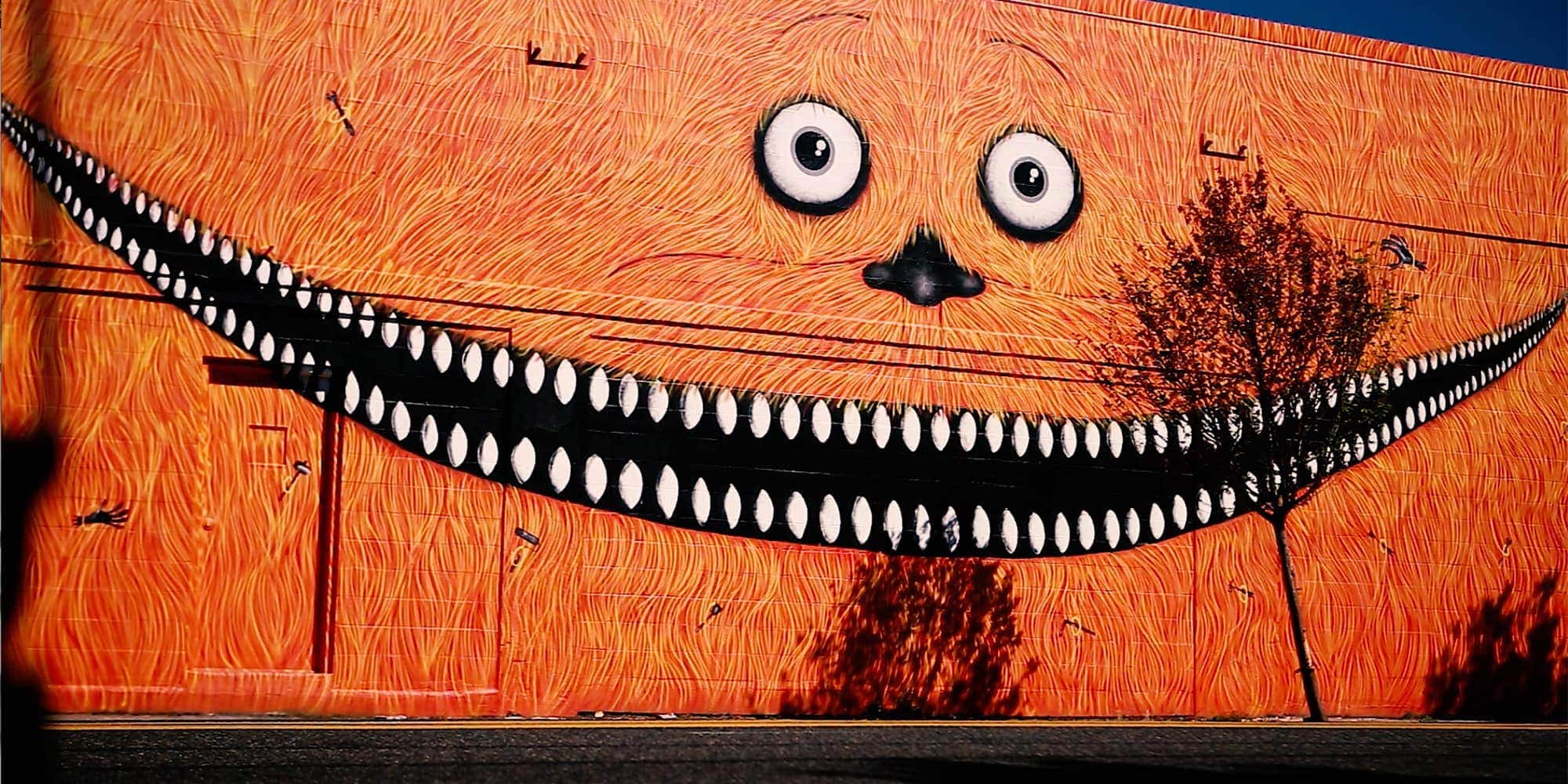 A mural with a wide, toothy grin and big eyes covers an orange, textured wall. A tree partially obscures the mouth, casting a shadow on the vibrant artwork.
