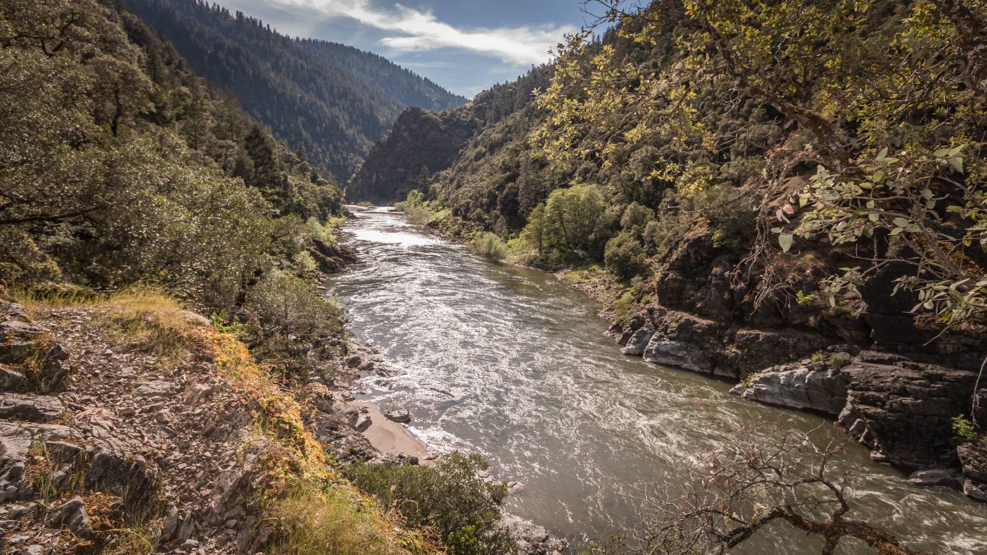 River flows through a narrow, tree-covered valley with rugged cliffs, under a partly cloudy sky, flanked by dense forests and distant mountains, creating a tranquil natural landscape.