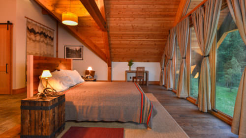A cozy bedroom features a rustic wooden bed with lamps on bedside tables. It overlooks a scenic view through large windows with draped curtains, under a sloped wooden ceiling.
