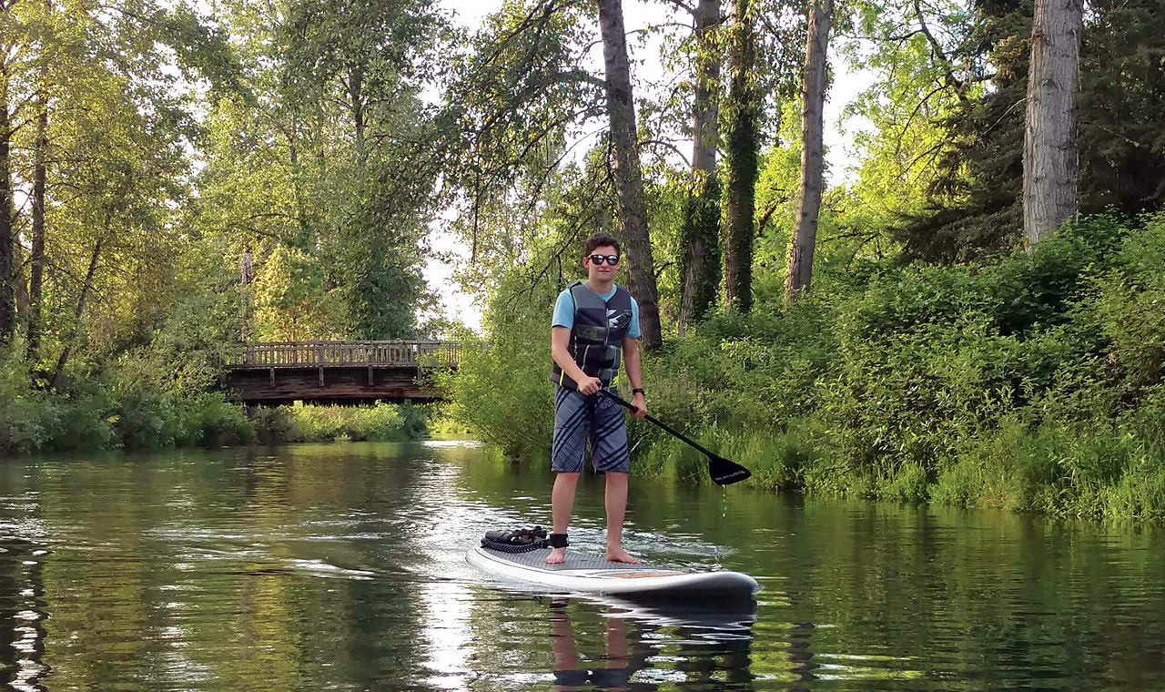 A boy paddles a board on a river surrounded by foliage
