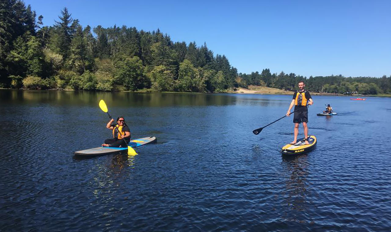 One person paddles a kayak and another paddles a stand-up paddleboard down a blue river
