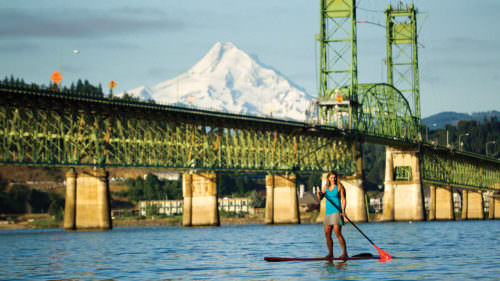 A woman holds a paddle on a stand-up paddleboard with bridge and mountain in background