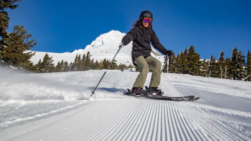 A skier wearing a helmet and goggles glides down a groomed snowy slope, holding poles. Tall pine trees and a snowy mountain are visible under a clear blue sky.