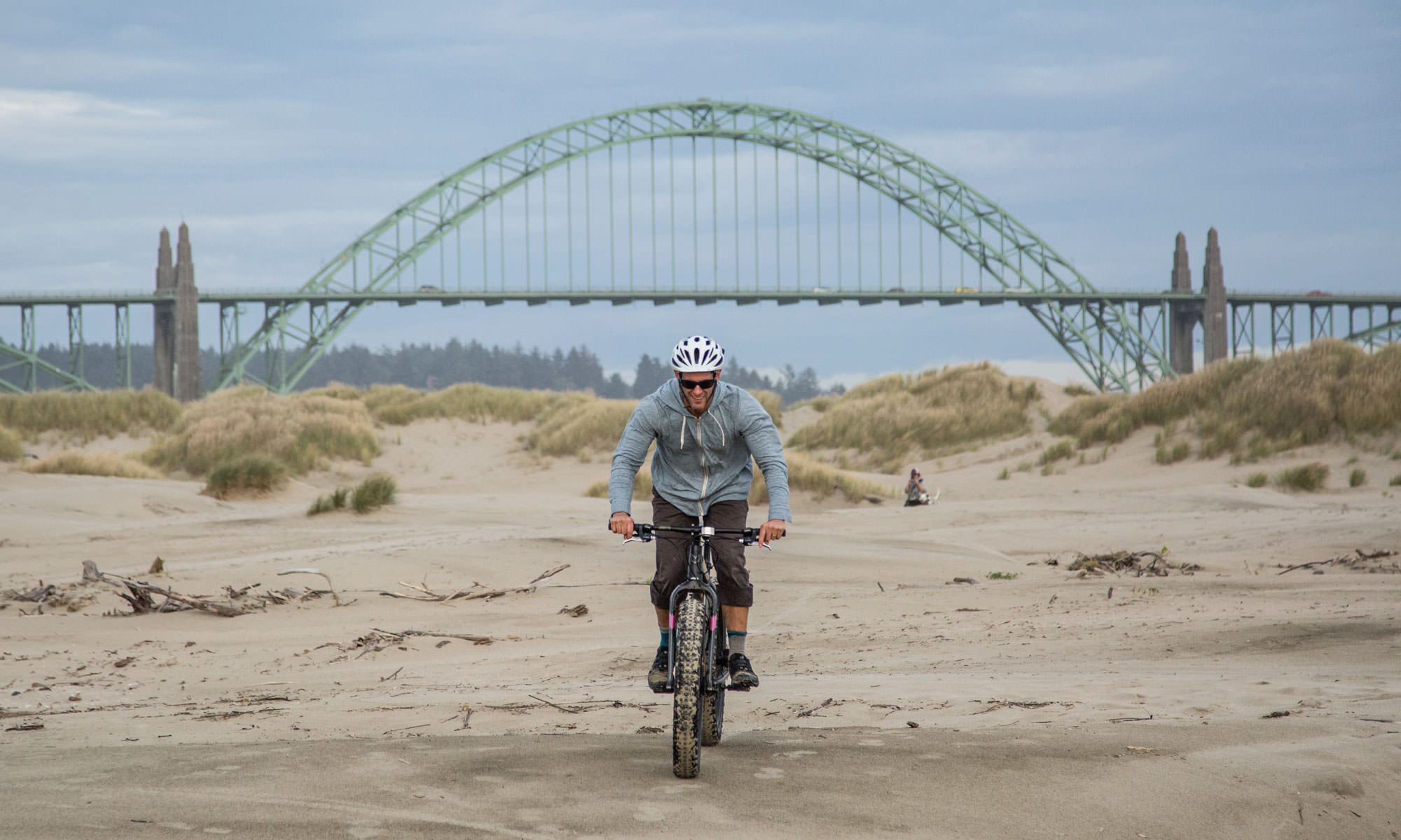 The green Newport Bridge looms in the background as a man gleefully rides a fat bike on the sand.