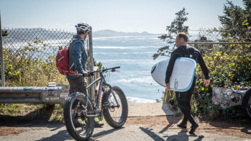 Man holding a fat bike shares the sidewalk with a surfer.