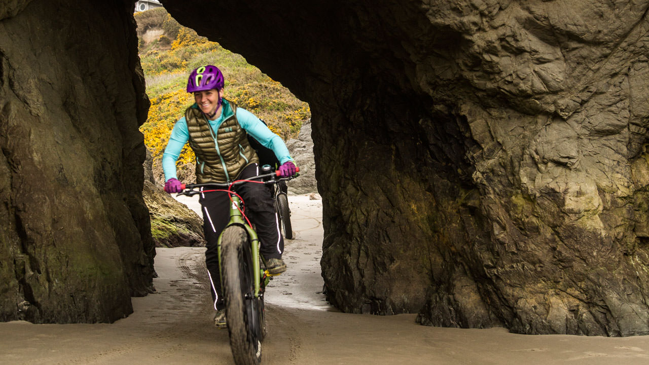 A woman pedals a fat-tire bike through an opening in a tall rock.