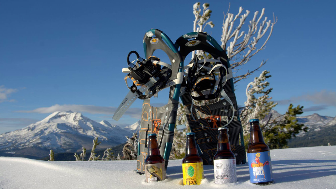 Four Central Oregon beer bottles sit in the snow, with two snowshoes behind them, as well as snowy peaks.