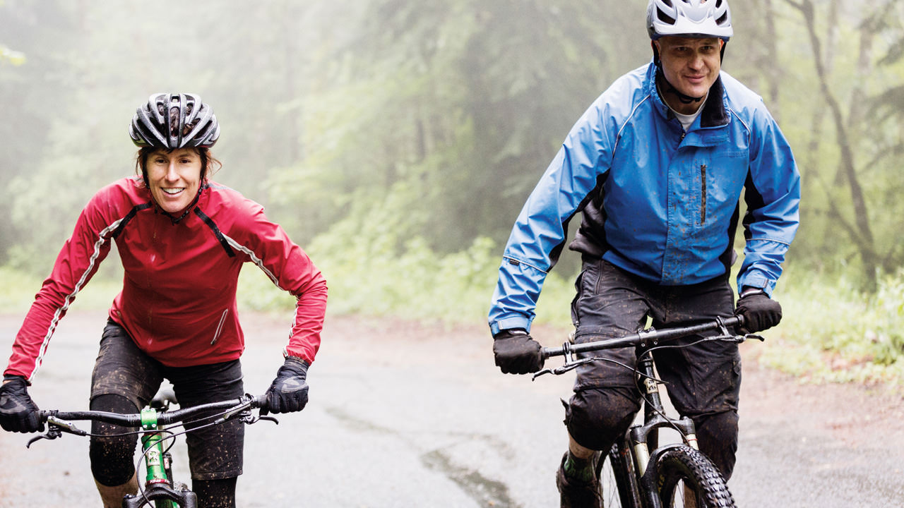 Two cyclists smile in the rain as they pedal up a hill in waterproof gear.