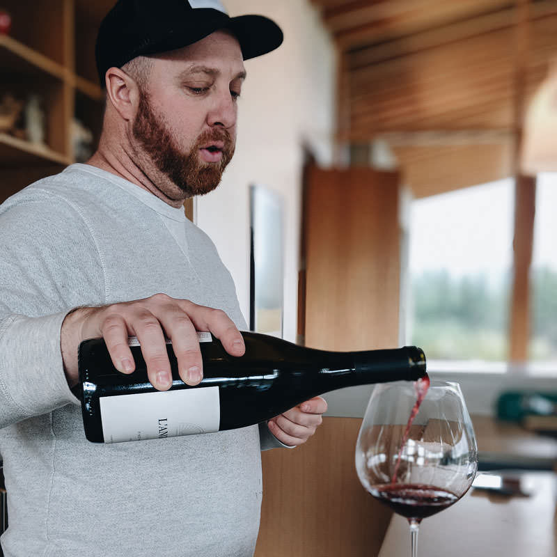A man wearing a gray sweatshirt delicately pours a rich pinot noir into a large wine glass.