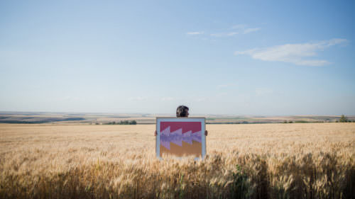 Person in a wheat field holding a brightly colored work of art and blue sky above.