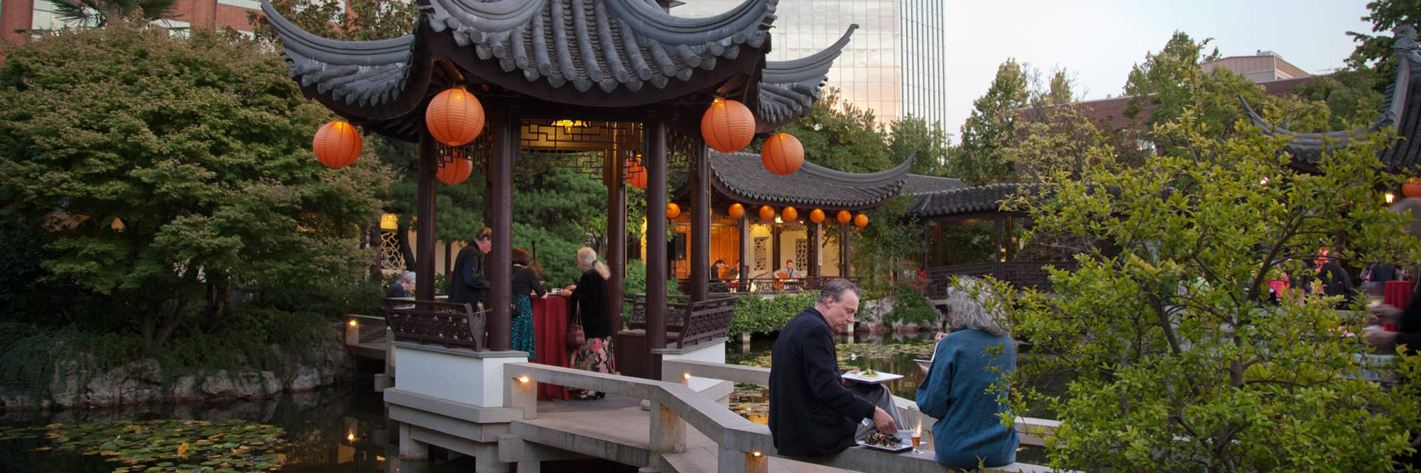 A traditional Chinese pavilion adorned with orange lanterns houses guests engaging in conversation and dining. The setting includes surrounding greenery, a reflective pond, and buildings in the background.