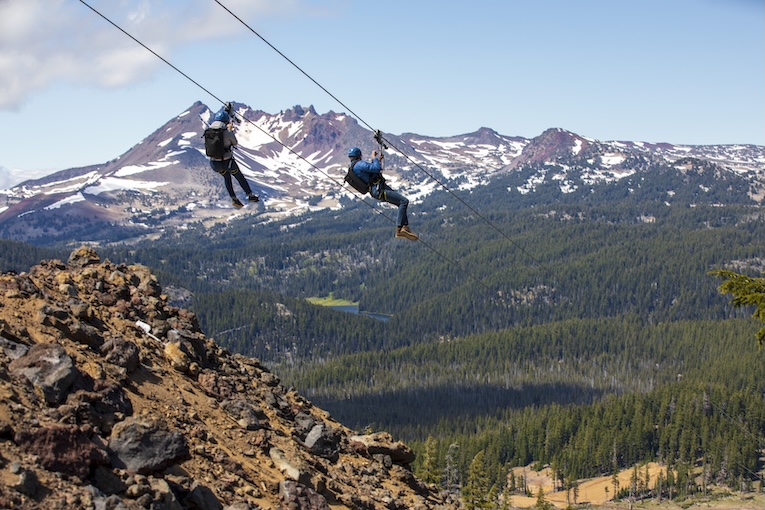 A pair of riders fly down the ziplin side-by-side on separate lines with mountain views. 