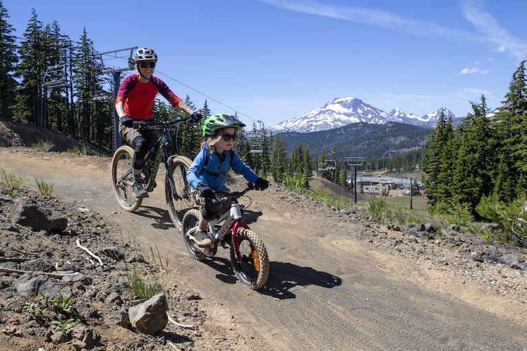  A toddler on a small strider bike leads the way down a dirt trail followed by an adult cyclist. 
