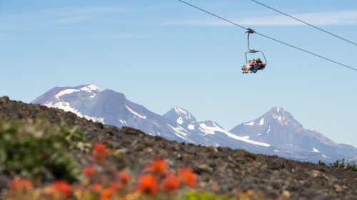 A chairlift carries several people over a rocky, flower-dotted landscape with snow-capped mountains in the background under a clear blue sky.