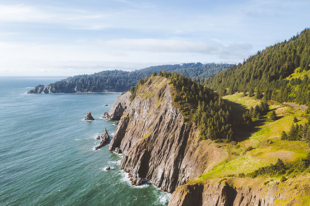 A bird's eye view of the Oregon Coast reveals lush landscape above rugged cliffs and blue ocean waters.