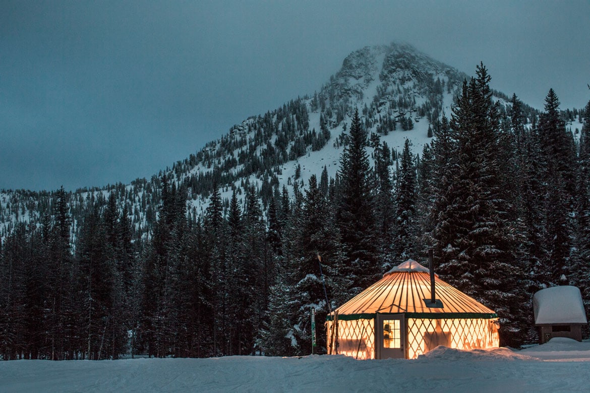 A yurt is illuminated against a snowy mountain backdrop near dusk.