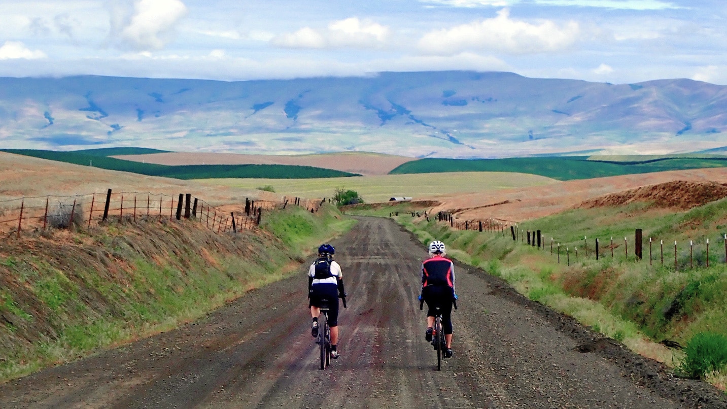 Two cyclists ride down a gravel road