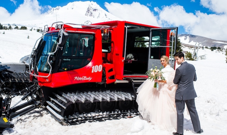 A bridge and groom stand in front of a snowcat.