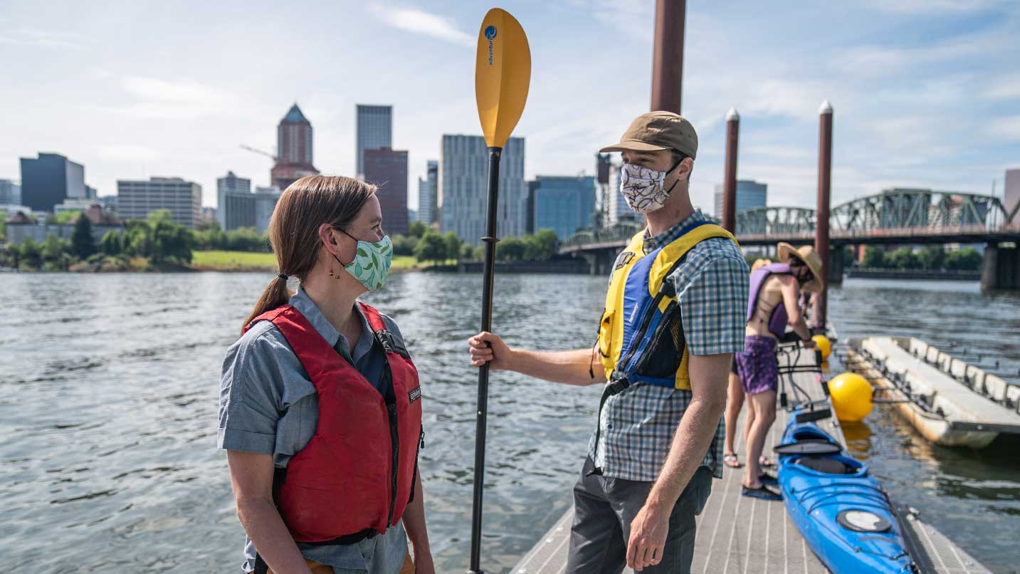 Two people prepare for kayaking while wearing life vests and face coverings.